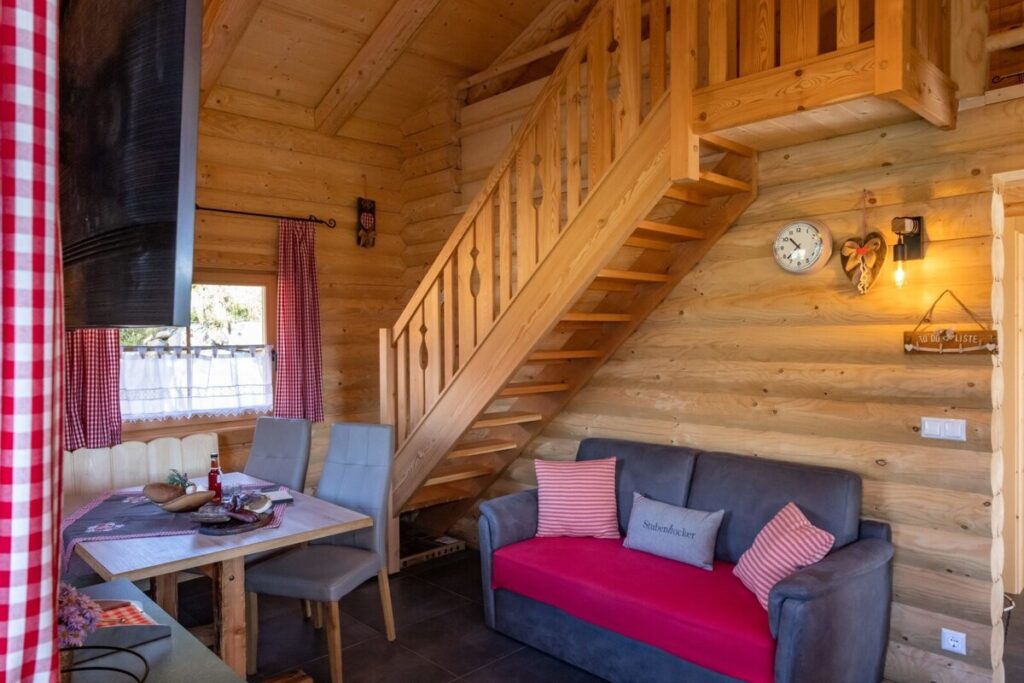 Der Wohn- und Essbereich der Alm mit Esstisch, Sofa und Holztreppe zum Obergeschoss.